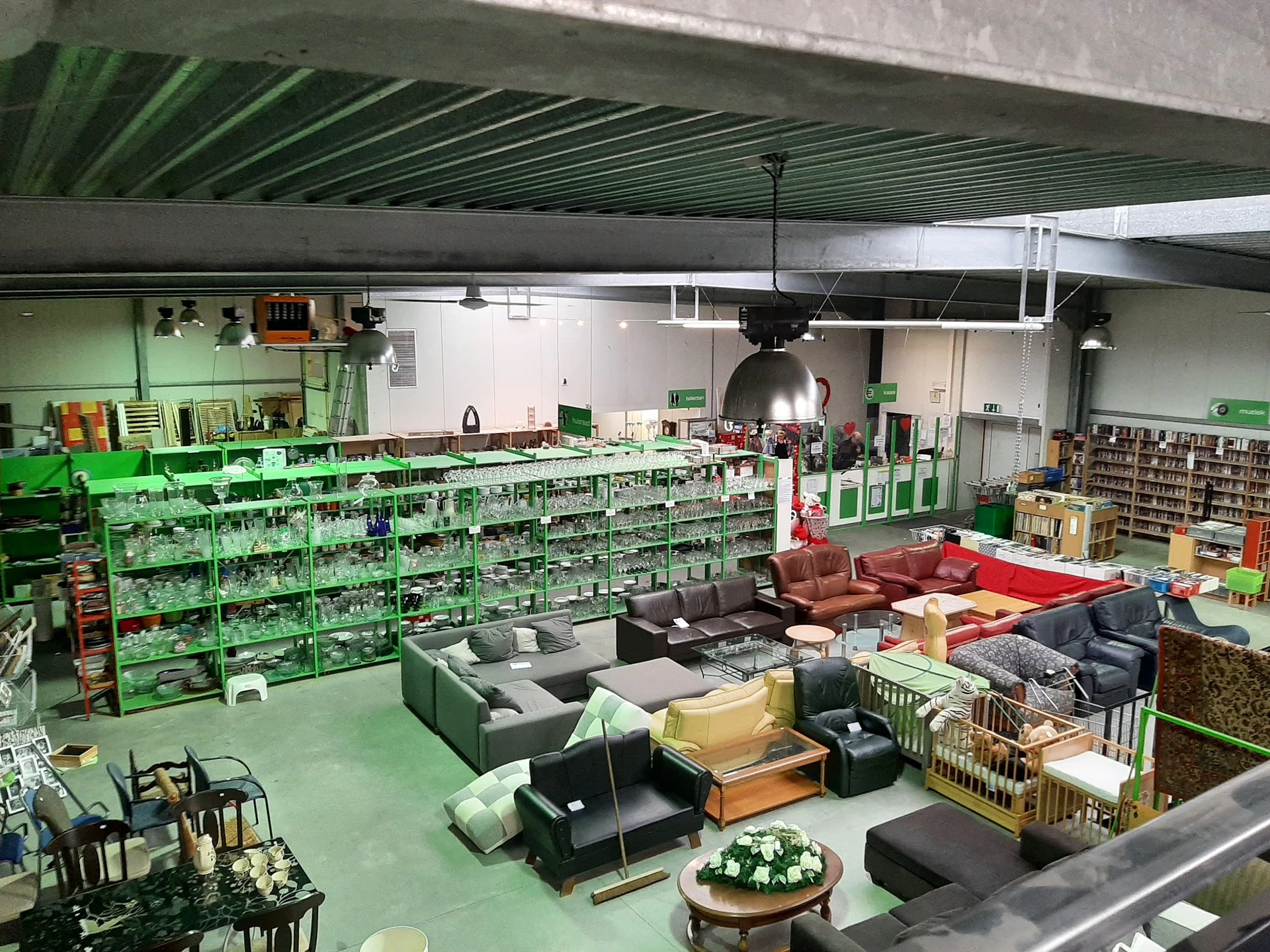 Overzicht van kringloopwinkelruimte met groene stellingen vol spullen, zitmeubels op de vloer en lichte productverlichting