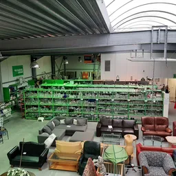 Ruim interieur van kringloopwinkel met groene stellingen vol spullen, zithoeken met banken en fauteuils op de vloer