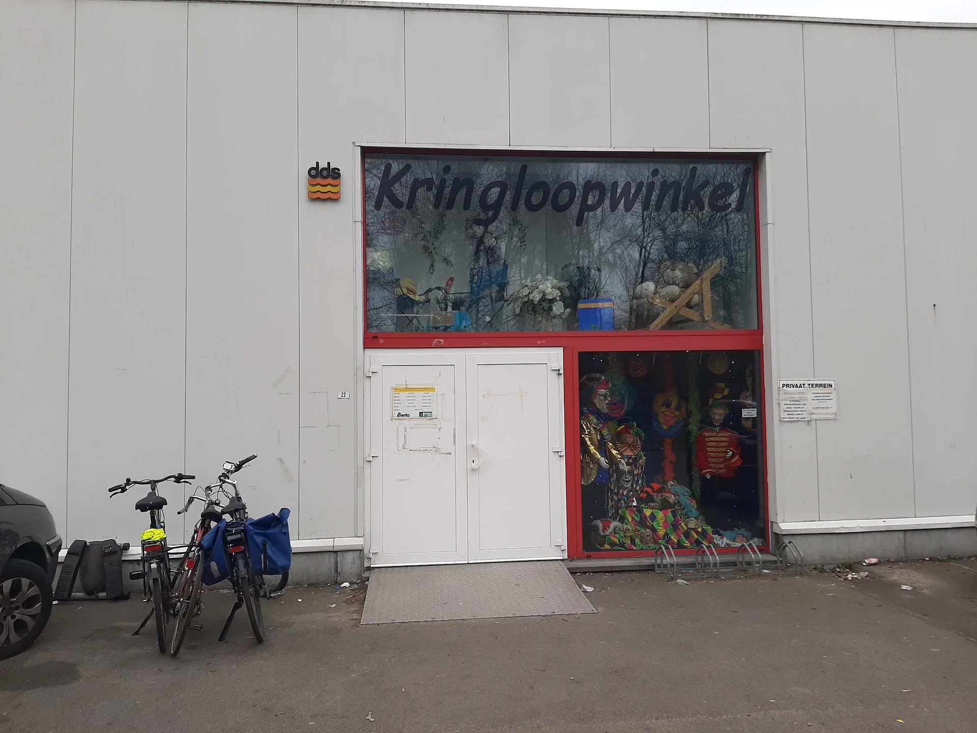 Gevel van kringloopwinkel met wit vlak en rode ingang, fietsen ervoor geparkeerd