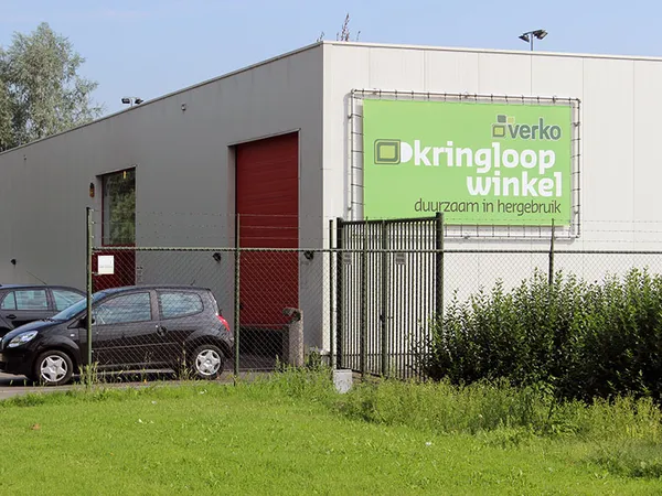 Grijze gevel van kringloopwinkel met groene reclamebanner 'Kringloopwinkel' en rode deur, parkeerplaats ervoor