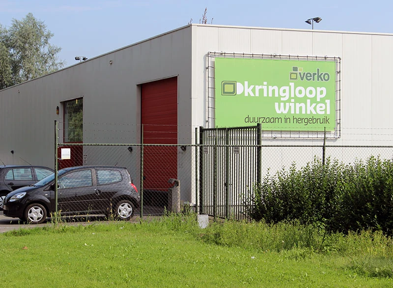 Grijze gevel van kringloopwinkel met groene reclamebanner 'Kringloopwinkel' en rode deur, parkeerplaats ervoor