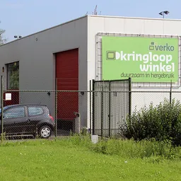 Grijze gevel van kringloopwinkel met groene reclamebanner 'Kringloopwinkel' en rode deur, parkeerplaats ervoor