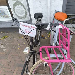 Roze kinderfietje met mandje en oranje zadelhoes, met handgeschreven verkoopbord 'Dames fiets 50,-'