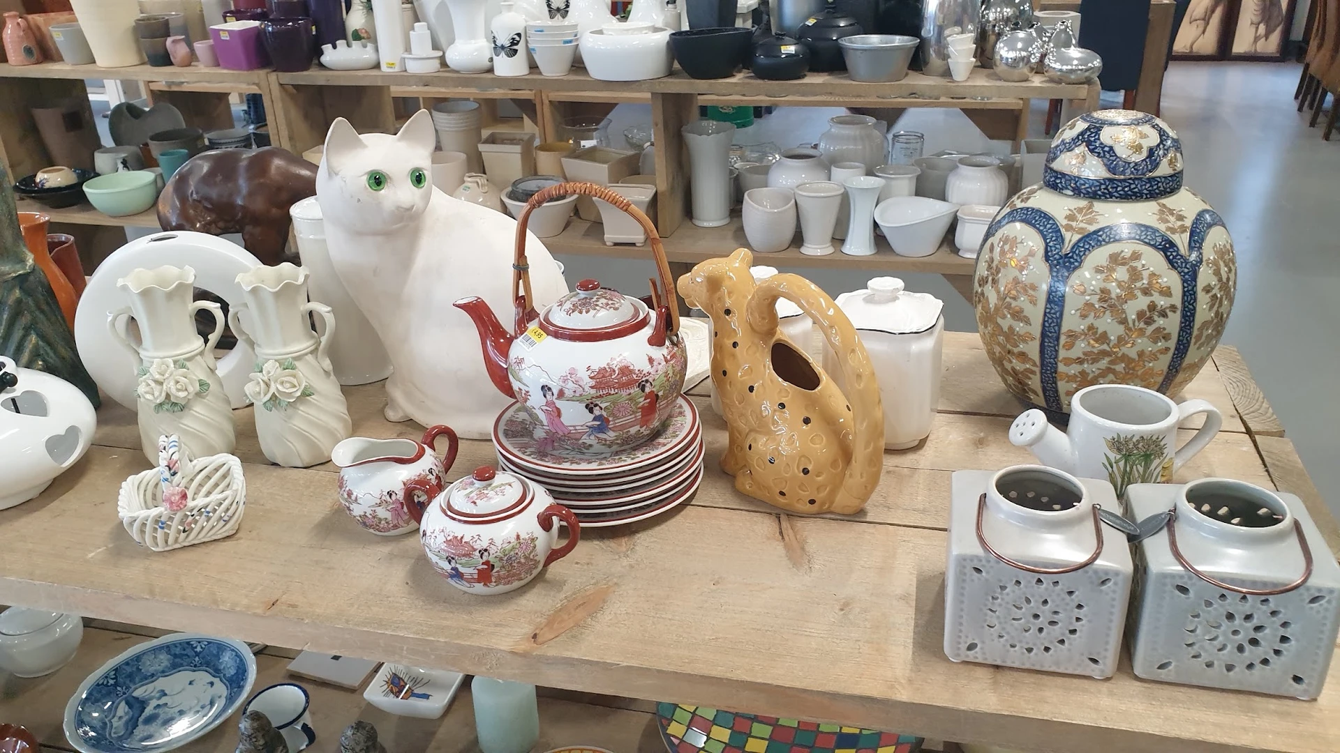Uitgestalde huishoudartikelen: witte kat, theepot met roze decoratie, borden, houten vaas en keramische items op tafel