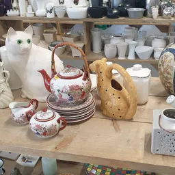 Uitgestalde huishoudartikelen: witte kat, theepot met roze decoratie, borden, houten vaas en keramische items op tafel