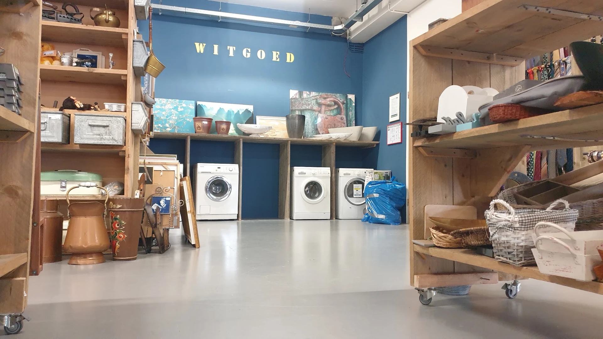 Witgoedafdeling met wasmachines onder houten stellingen en planken met huishoudartikelen