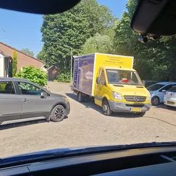 Gele bedrijfsbus met logo van kringloopwinkel geparkeerd op werkterrein tussen andere auto's