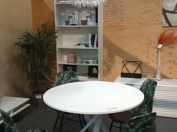 Winkelinterieur met ronde witte tafel, groene stoelen, witte stellingkast, planten en houten muren