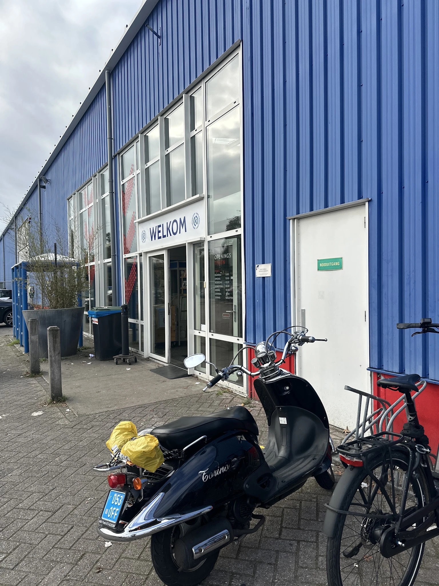 Ingang van kringloopwinkel met WELKOM-bord, blauwe golfplaten wand, scooters en fietsen op parkeerplaats