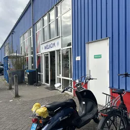 Ingang van kringloopwinkel met WELKOM-bord, blauwe golfplaten wand, scooters en fietsen op parkeerplaats