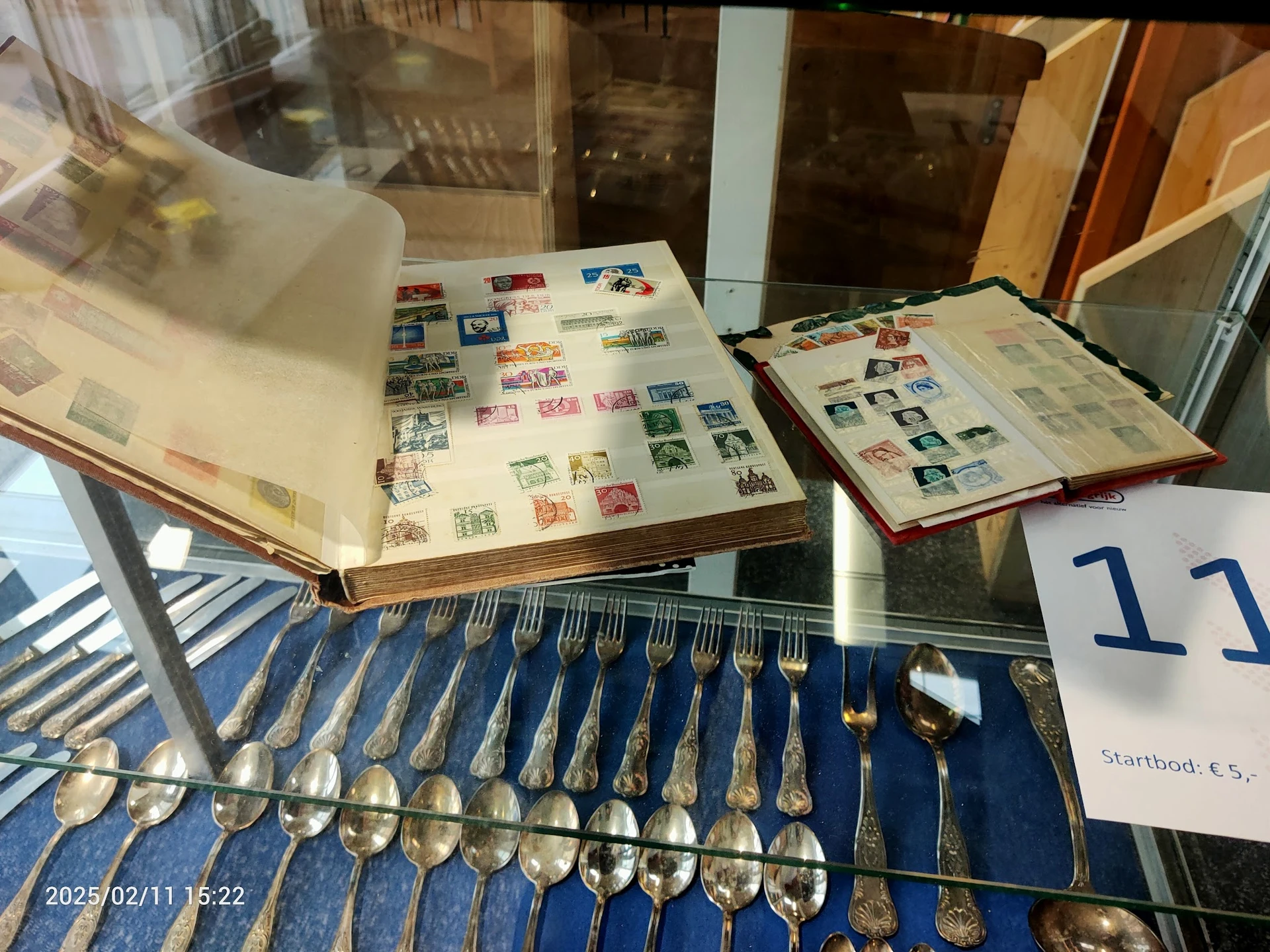 Vitrinekast met verzamelde postzegels in albums en een set zilveren lepels en vorken op blauwe achtergrond