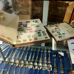 Vitrinekast met verzamelde postzegels in albums en een set zilveren lepels en vorken op blauwe achtergrond