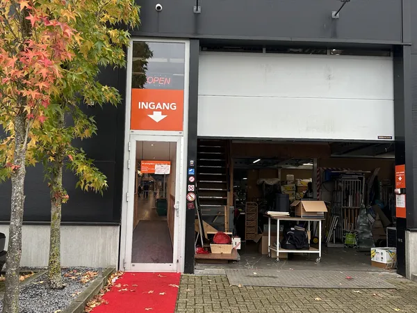 Voorkant kringloopwinkel met oranje INGANG-bord, uitstalramen met meubels en spullen, rode mat en herfstbladeren