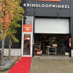Voorkant kringloopwinkel met oranje INGANG-bord, uitstalramen met meubels en spullen, rode mat en herfstbladeren