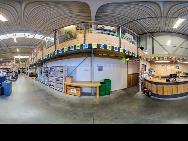 Ruim interieur van kringloopwinkel met houten plafond, stellingen, informatieborden en balie aan rechterkant