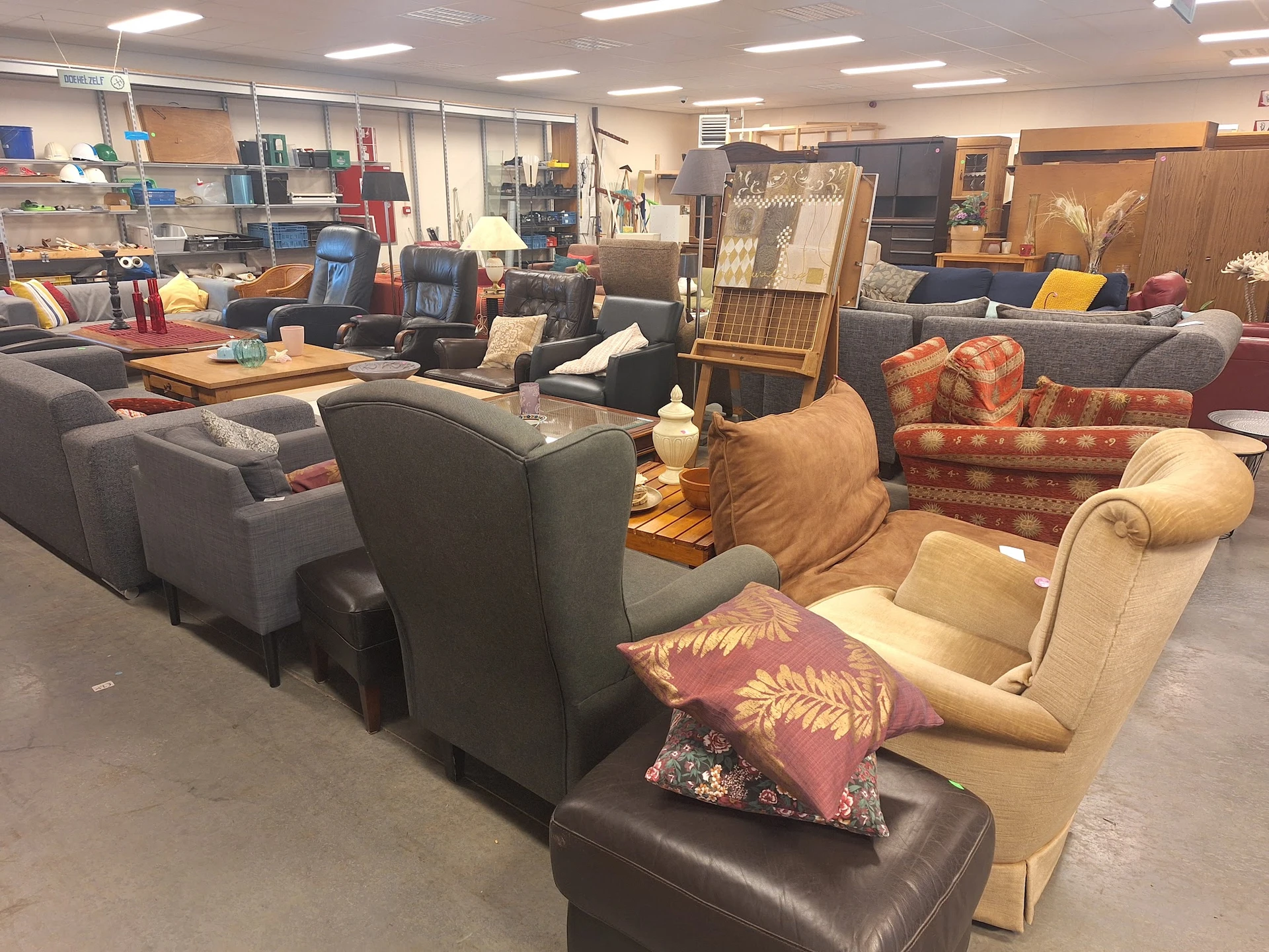 Winkelvloer met diverse zitmeubels: grijze stoelen, beige fauteuils, rode pluche bank en andere tweedehands meubelstukken uit
