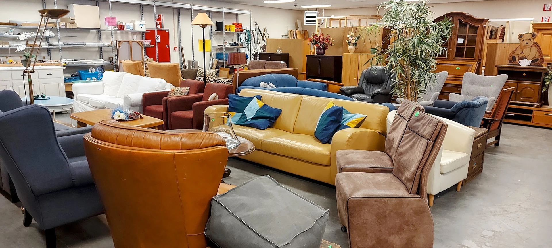 Diverse tweedehands meubels: gele bank met blauwe kussens, bruine leren stoelen en fauteuils in de kringloopwinkel