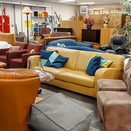 Diverse tweedehands meubels: gele bank met blauwe kussens, bruine leren stoelen en fauteuils in de kringloopwinkel