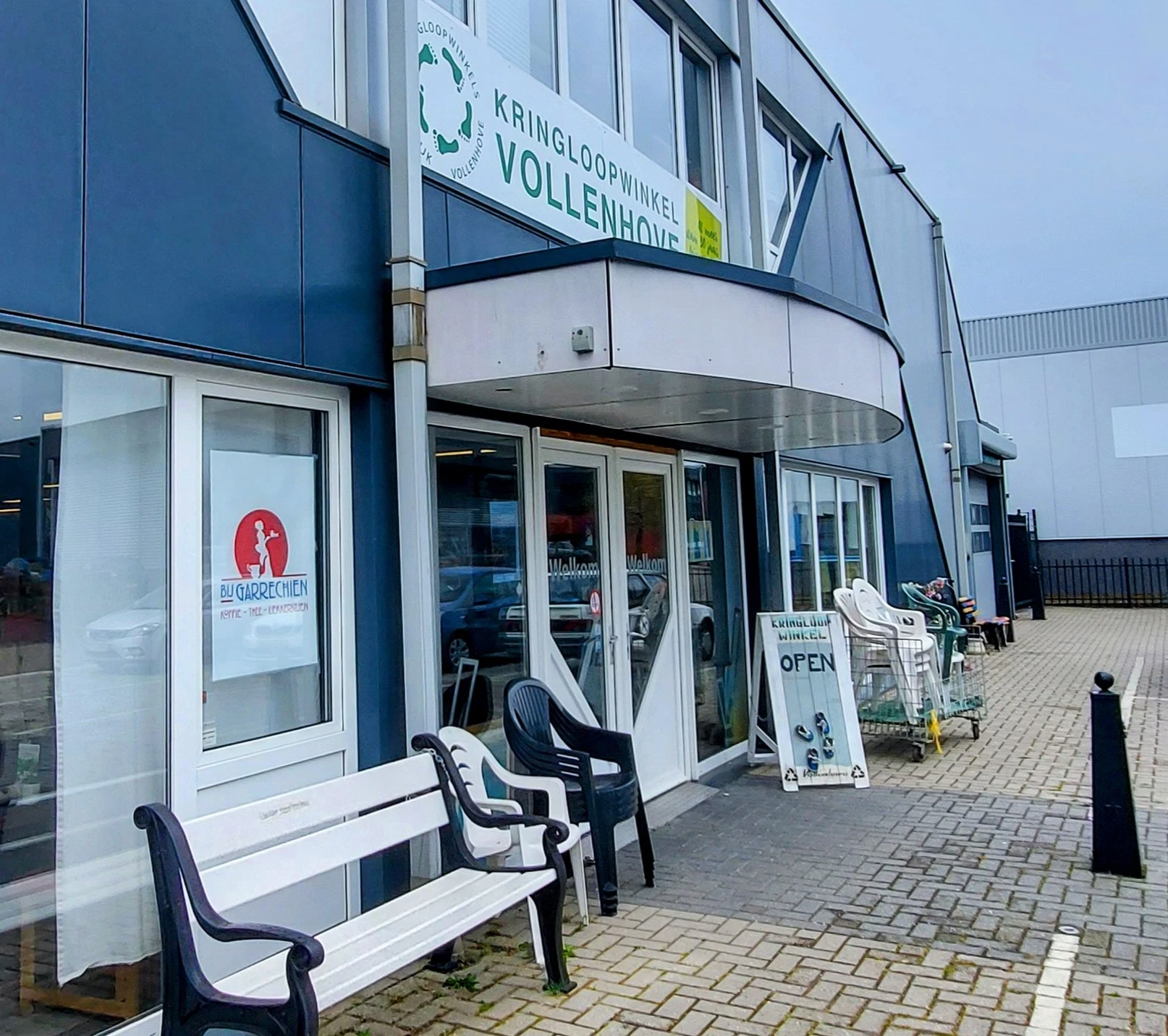 Moderne gevel van kringloopwinkel met glazen deuren, blauwe en witte bekleding, terras met stoelen en openlucht uitstalling