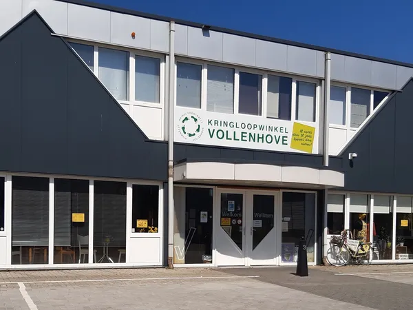Gevel van kringloopwinkel Vollenhove met naam op witte band, zwarte zijgevels, grote ramen en ingang met donkere deuren.