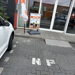 Winkelvoorgevel met witte deuren en welkomstbord, parkeerplek met witte auto linksvoor
