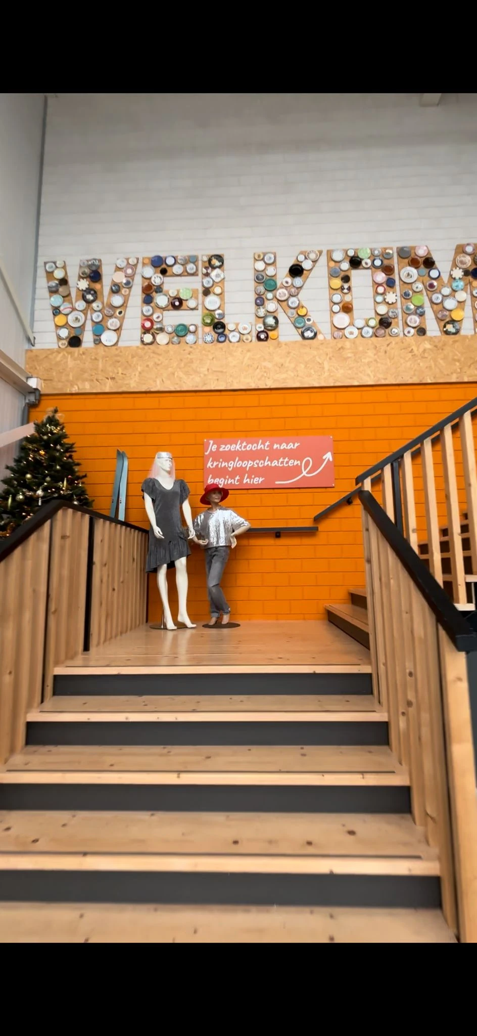 Trap met houten leuning in kringloopwinkel, pop-up etalage met kledingpop en rood bord met Nederlandse tekst op oranje muur
