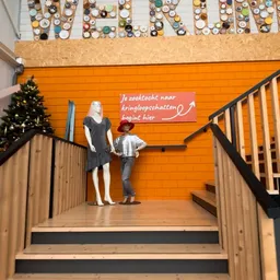 Trap met houten leuning in kringloopwinkel, pop-up etalage met kledingpop en rood bord met Nederlandse tekst op oranje muur