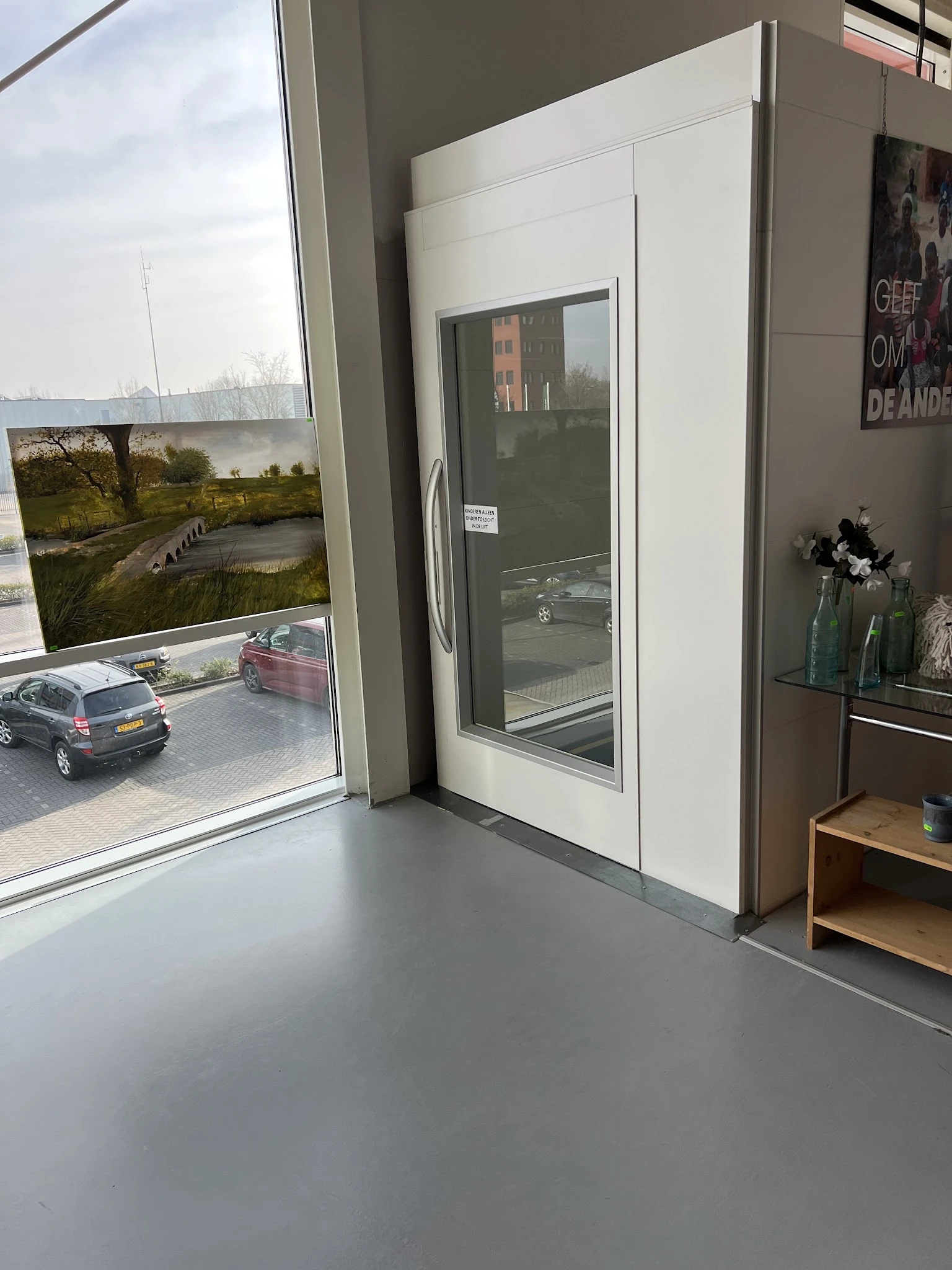 Winkelinterieur met grote ramen, uitzicht op parkeerplaats en tuin, stellingen met spullen aan rechterkant