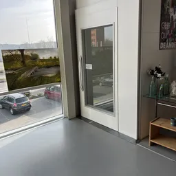 Winkelinterieur met grote ramen, uitzicht op parkeerplaats en tuin, stellingen met spullen aan rechterkant