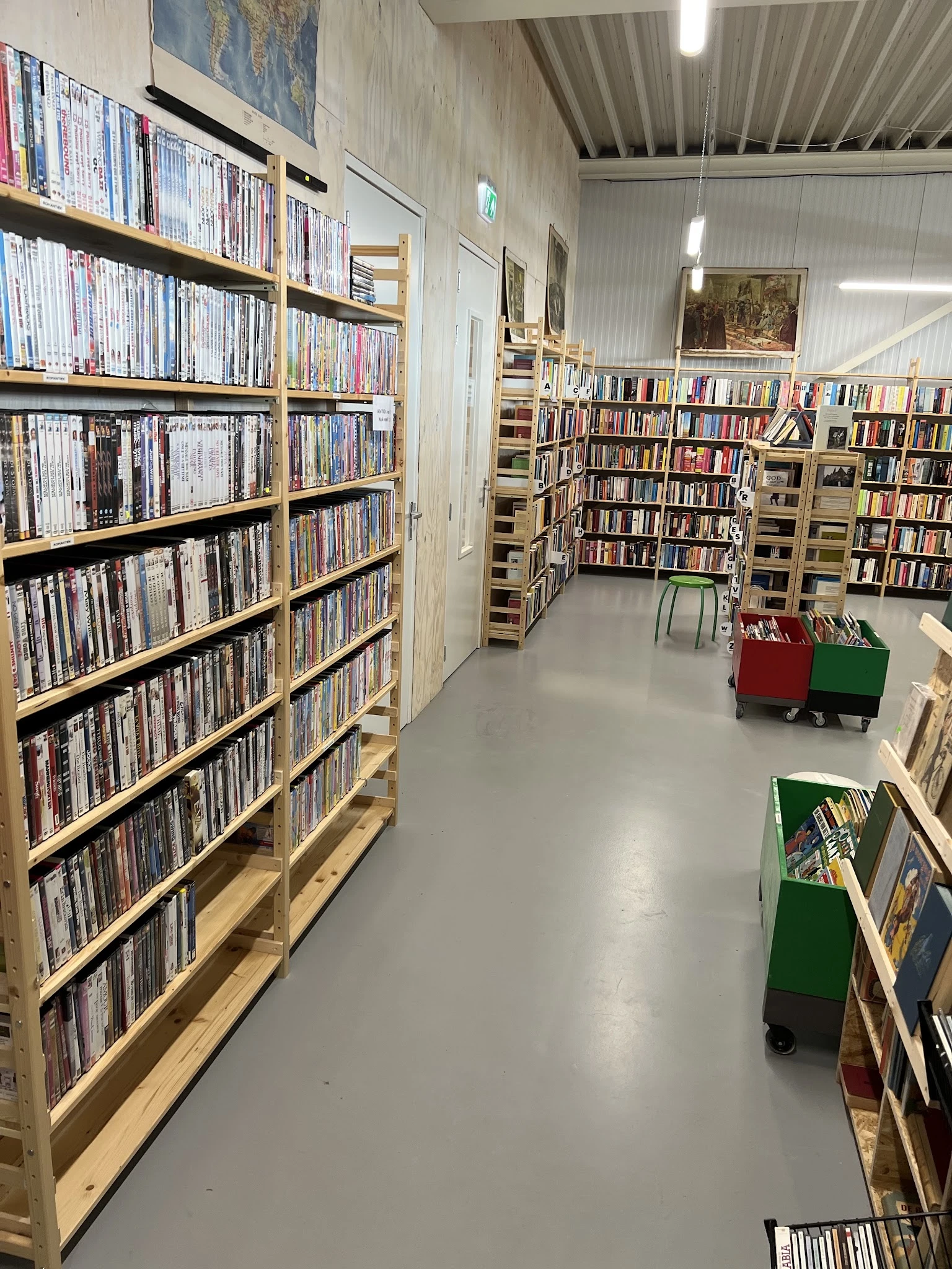 Binnenaanzicht van kringloopwinkel met houten stellingen vol dvd's en boeken, groene en rode kratjes op wielen op de grond