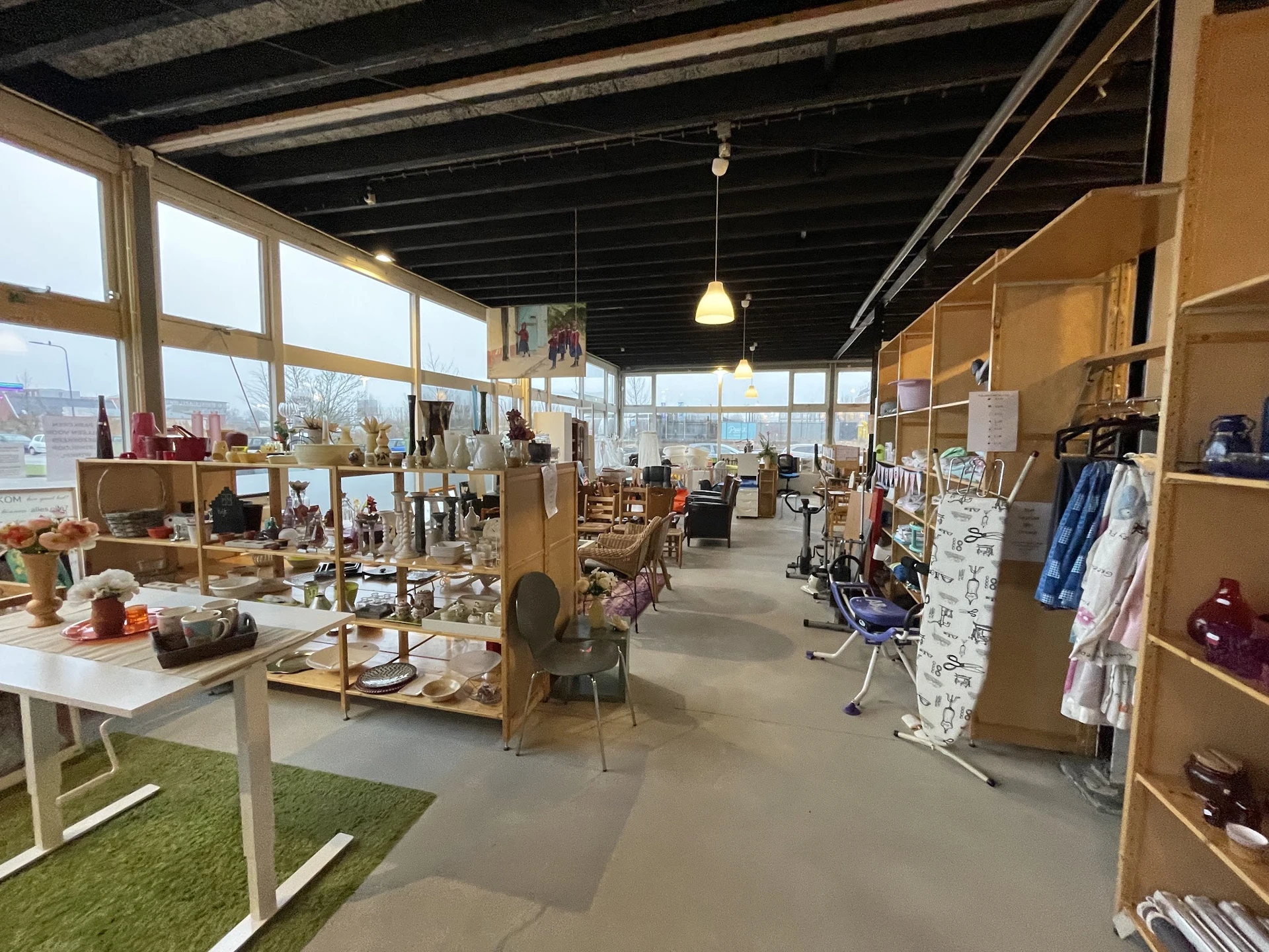 Ruim winkelinterieur met houten stellingen, kledingrekken, huishoudelijke spullen en een groen vloergedeelte, grote ramen met