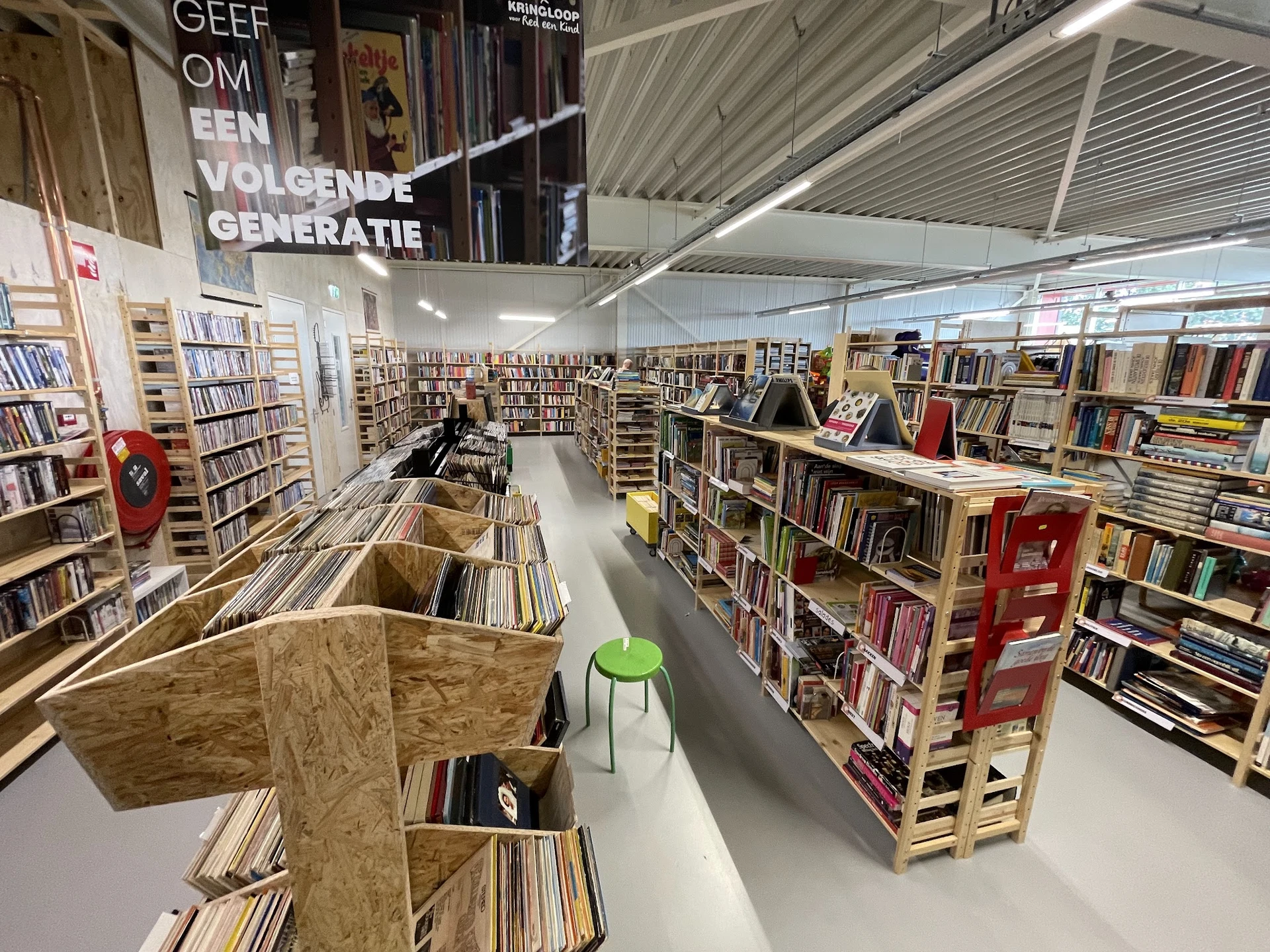 Ruime winkelruimte met kasten en stellingen vol cd's, dvd's en boeken, groen stoeltje zichtbaar