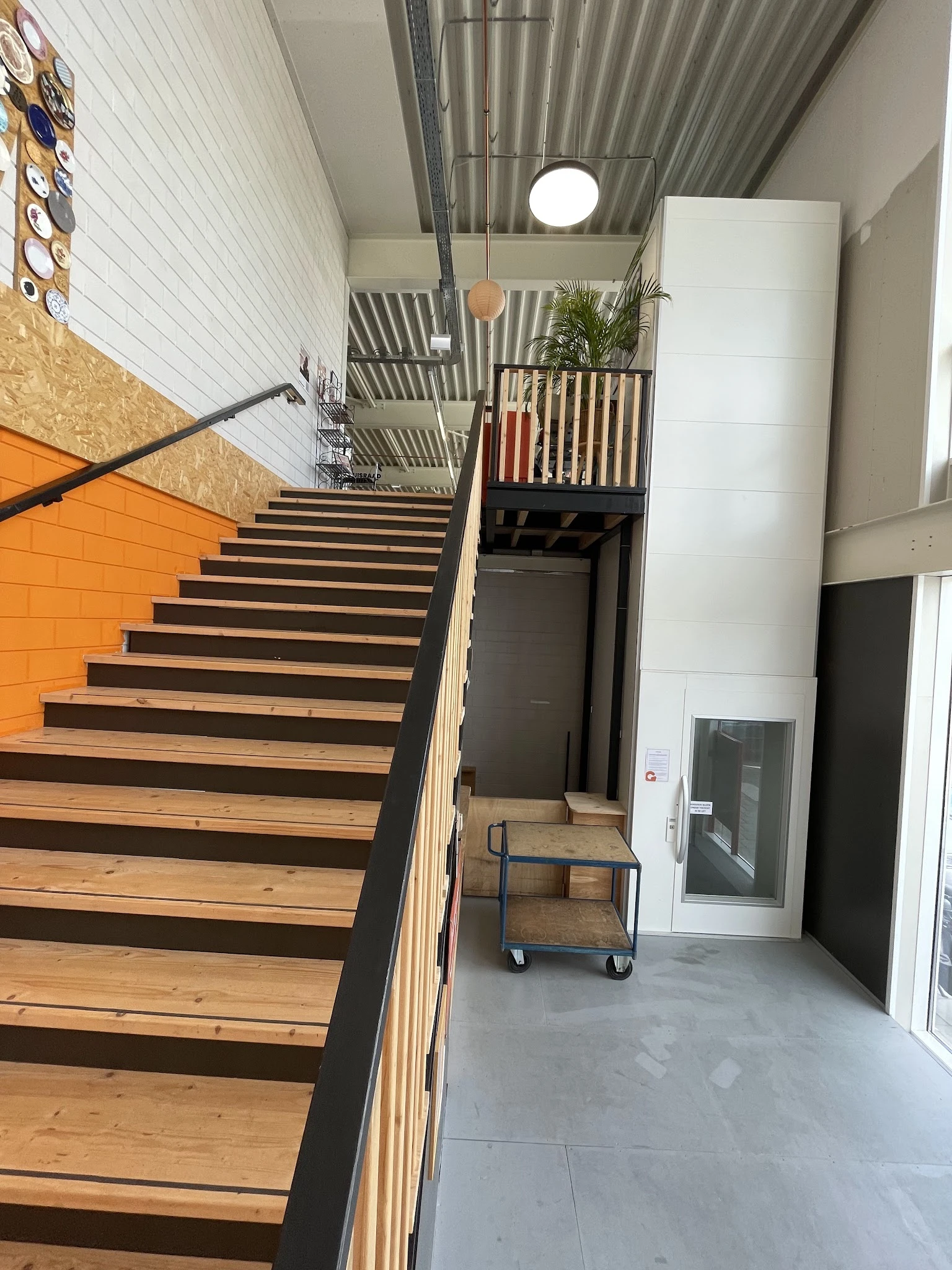 Binnenaanzicht van kringloopwinkel met houten trap, zwarte trapleuning, oranje muur, stellingen en gang naar andere ruimtes