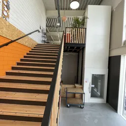 Binnenaanzicht van kringloopwinkel met houten trap, zwarte trapleuning, oranje muur, stellingen en gang naar andere ruimtes