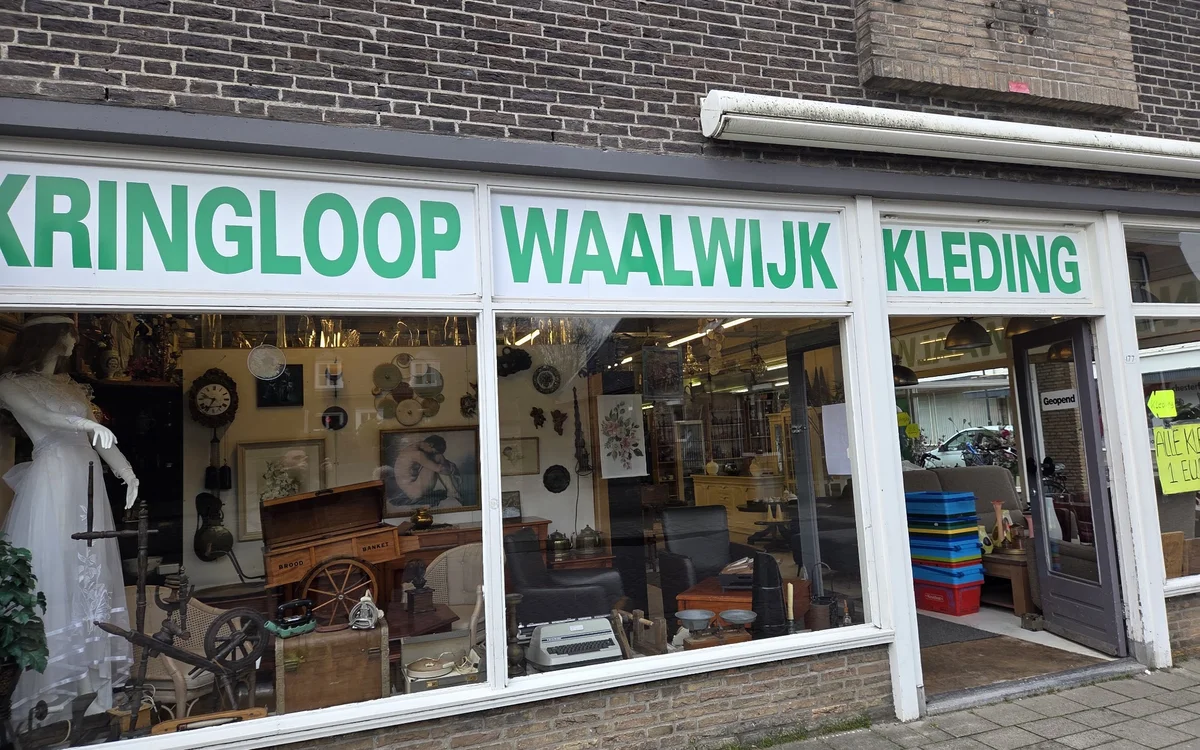 Winkelgevel met drie grote ramen, witte raamkozijnen en groene letters. Zichtbaar zijn verschillende tweedehands spullen in d