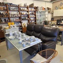 Winkelvloer met zwarte leren bank, tafels met servies en blauw-wit aardewerk, stellingen vol collectibles en decoratie