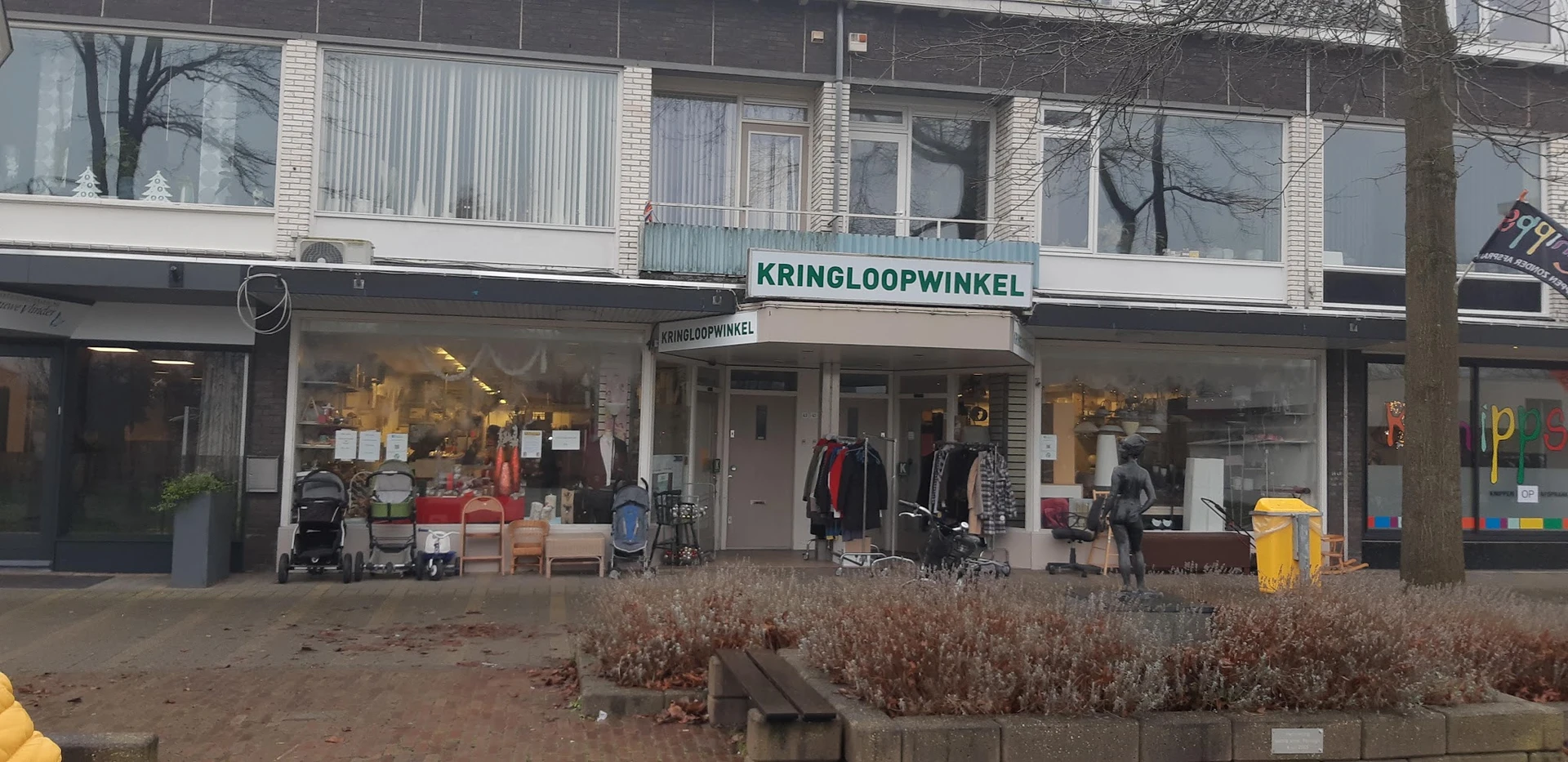 Gevel van kringloopwinkel met winkelnaam KRINGLOOPWINKEL op de pui, etalage met spullen en kleding zichtbaar
