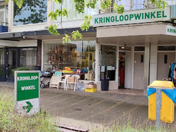 Voorkant van kringloopwinkel met groene gevel, uitgestalde spullen buiten, groene en gele prullenbakken.