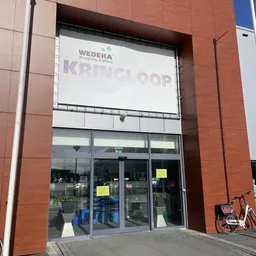 Gevel van kringloopwinkel Wedeka met grote glazen ingang en witte gevel, winkelnaam zichtbaar op bord