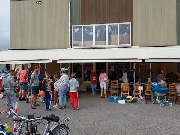 Voorkant kringloopwinkel met open deuren, bezoekers en uitgestalde spullen op het terras