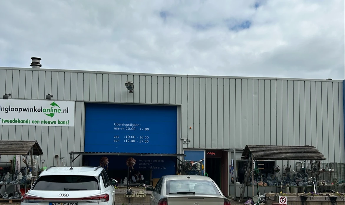 Grijze metalen gevel van kringloopwinkel met blauwe deur en openingstijden, geparkeerde auto's ervoor