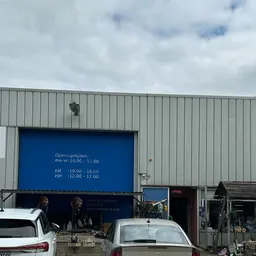 Grijze metalen gevel van kringloopwinkel met blauwe deur en openingstijden, geparkeerde auto's ervoor