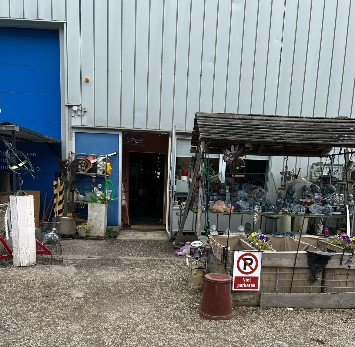 Voorkant van kringloopwinkel met metalen gevel, ingang en uitstallingen met bloemen en voorwerpen buiten