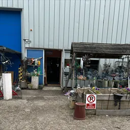 Voorkant van kringloopwinkel met metalen gevel, ingang en uitstallingen met bloemen en voorwerpen buiten