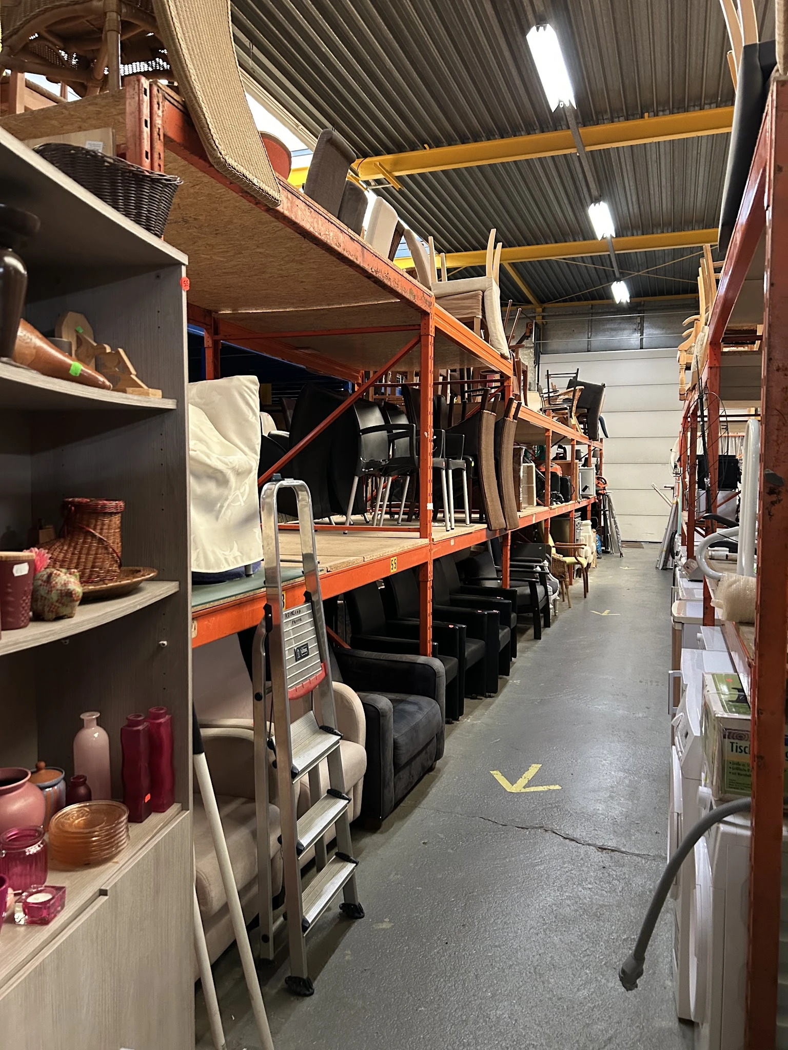 Magazijngang met oranje stellingen vol opgeslagen meubels en huishoudartikelen, linker wand met zwarte kasten en decoratieve 