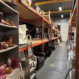 Magazijngang met oranje stellingen vol opgeslagen meubels en huishoudartikelen, linker wand met zwarte kasten en decoratieve 