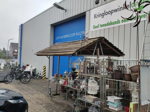 Voorkant kringloopwinkel met blauwe deuren, winkelnaam op gevel, uitgestalde spullen onder afdak