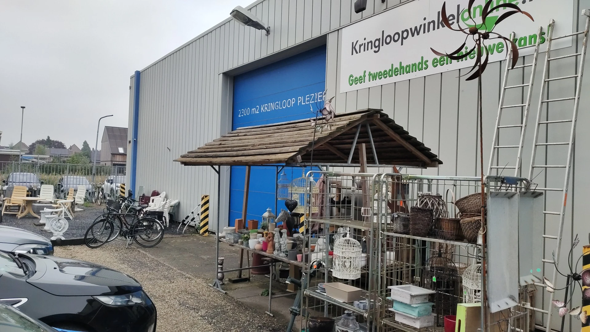 Voorkant kringloopwinkel met blauwe deuren, winkelnaam op gevel, uitgestalde spullen onder afdak