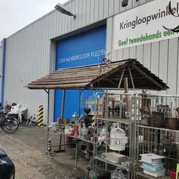 Voorkant kringloopwinkel met blauwe deuren, winkelnaam op gevel, uitgestalde spullen onder afdak