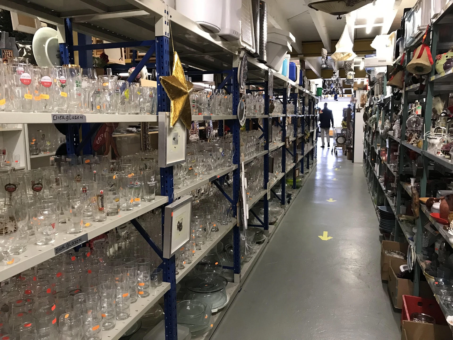 Lange rijen stellingen met honderden glazen drinkglazen en servies uitgestald in een kringloopwinkel